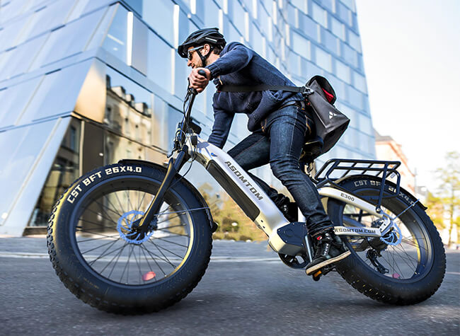 ASOMTOM electric bike in action, rider wearing a helmet on the E300 model with fat tires, ideal for urban and off-road use, against a modern building backdrop